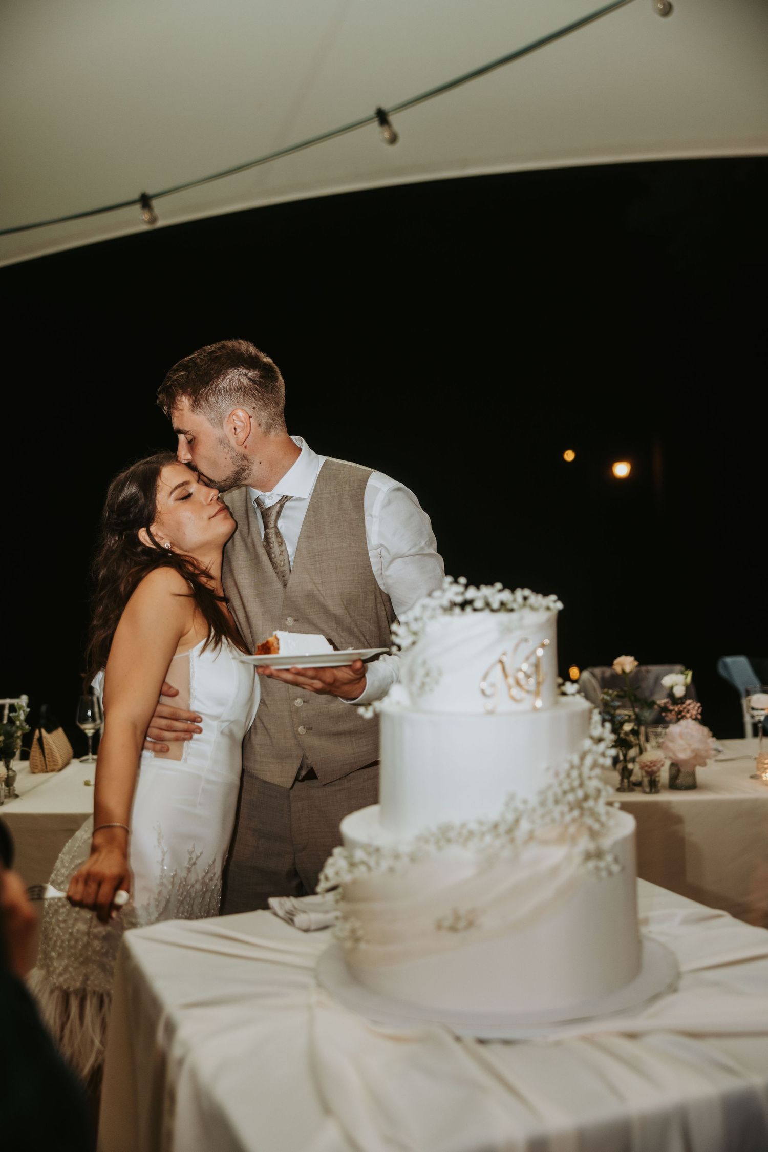 Bride and groom share a kiss while cutting their elegant three-tier white wedding cake at reception.