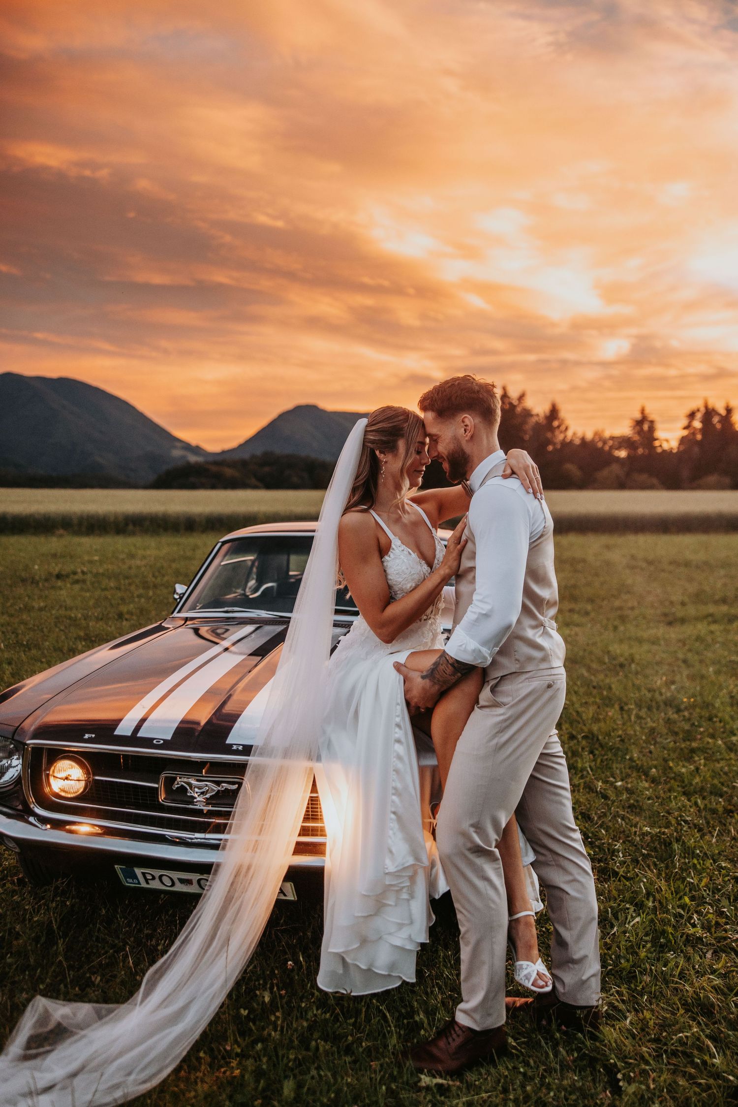 Bride and groom share romantic embrace leaning against classic Mustang at vibrant sunset.