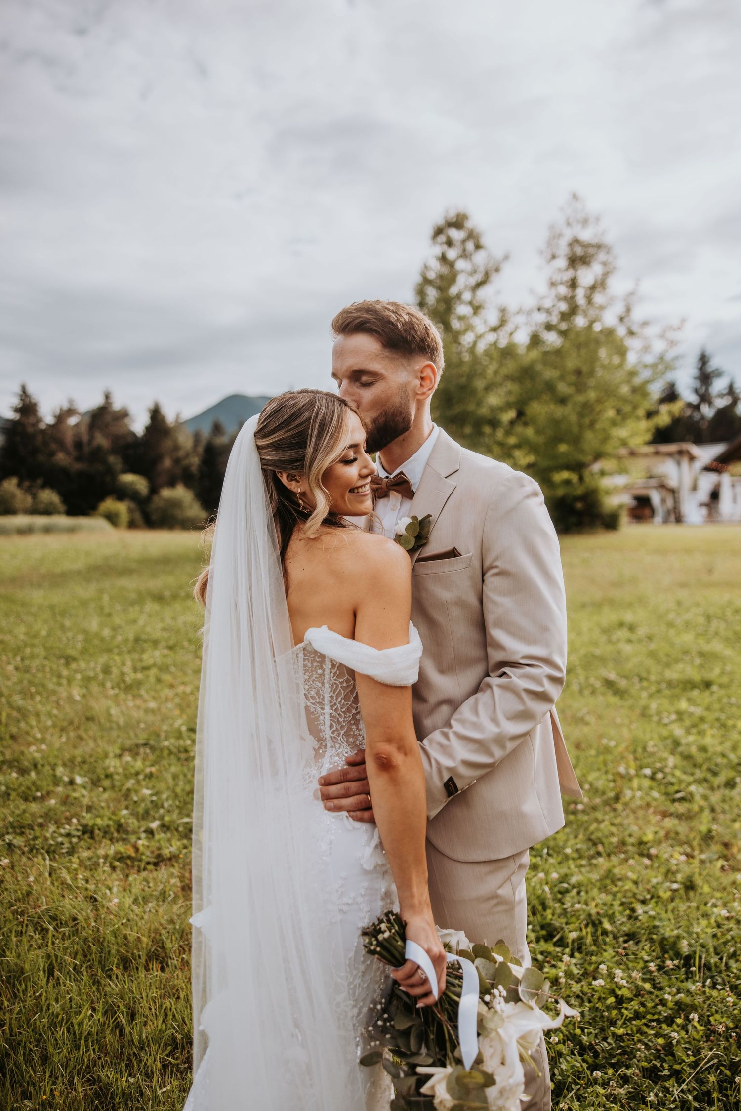 Groom in beige suit kisses bride in off-shoulder lace gown in a green meadow with mountain backdrop.