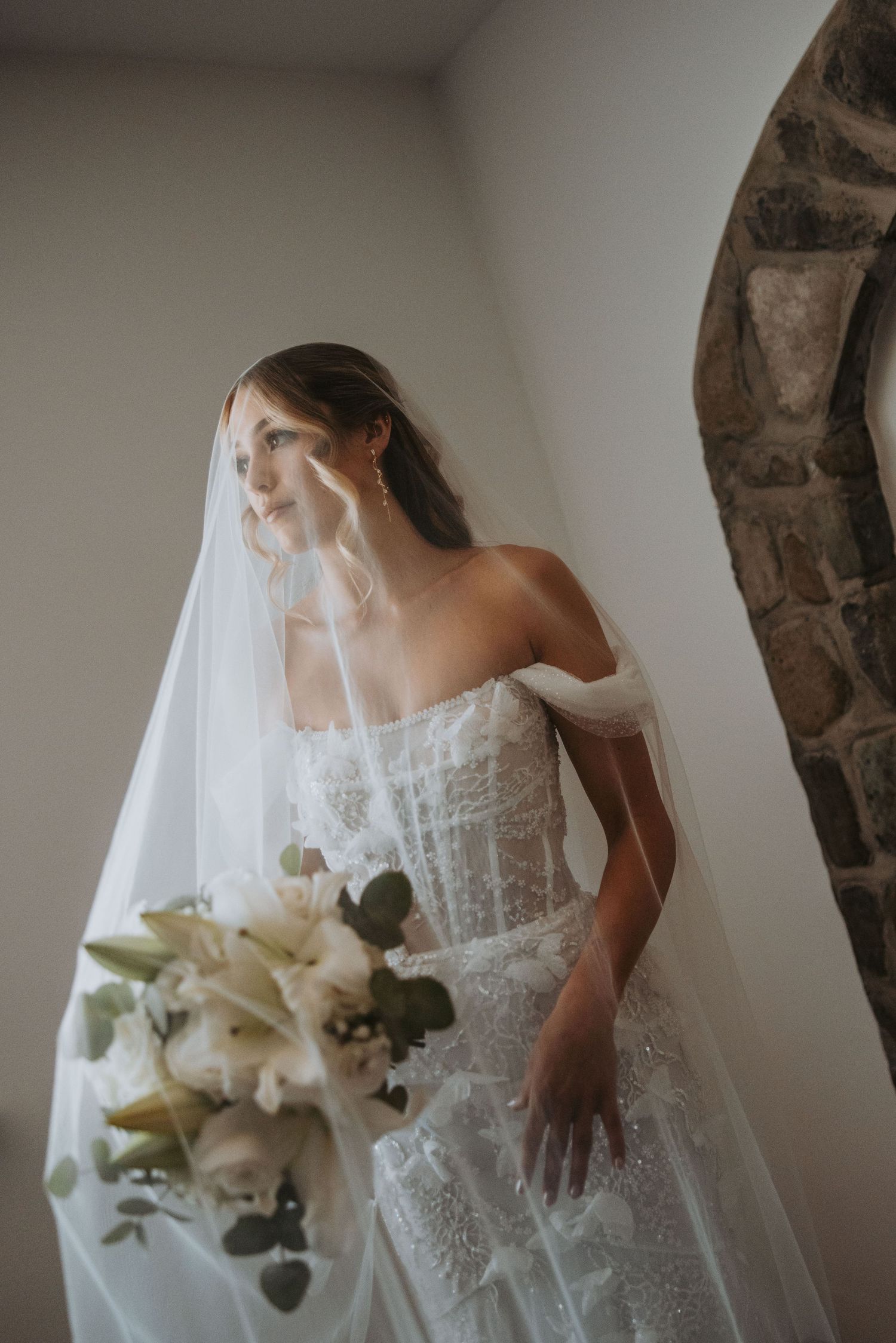 Beautiful bride in off-shoulder lace wedding gown and veil holds white lily bouquet by stone arch.