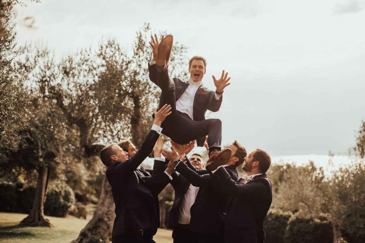 Groomsmen tossing groom in the air outdoors during wedding celebration.
