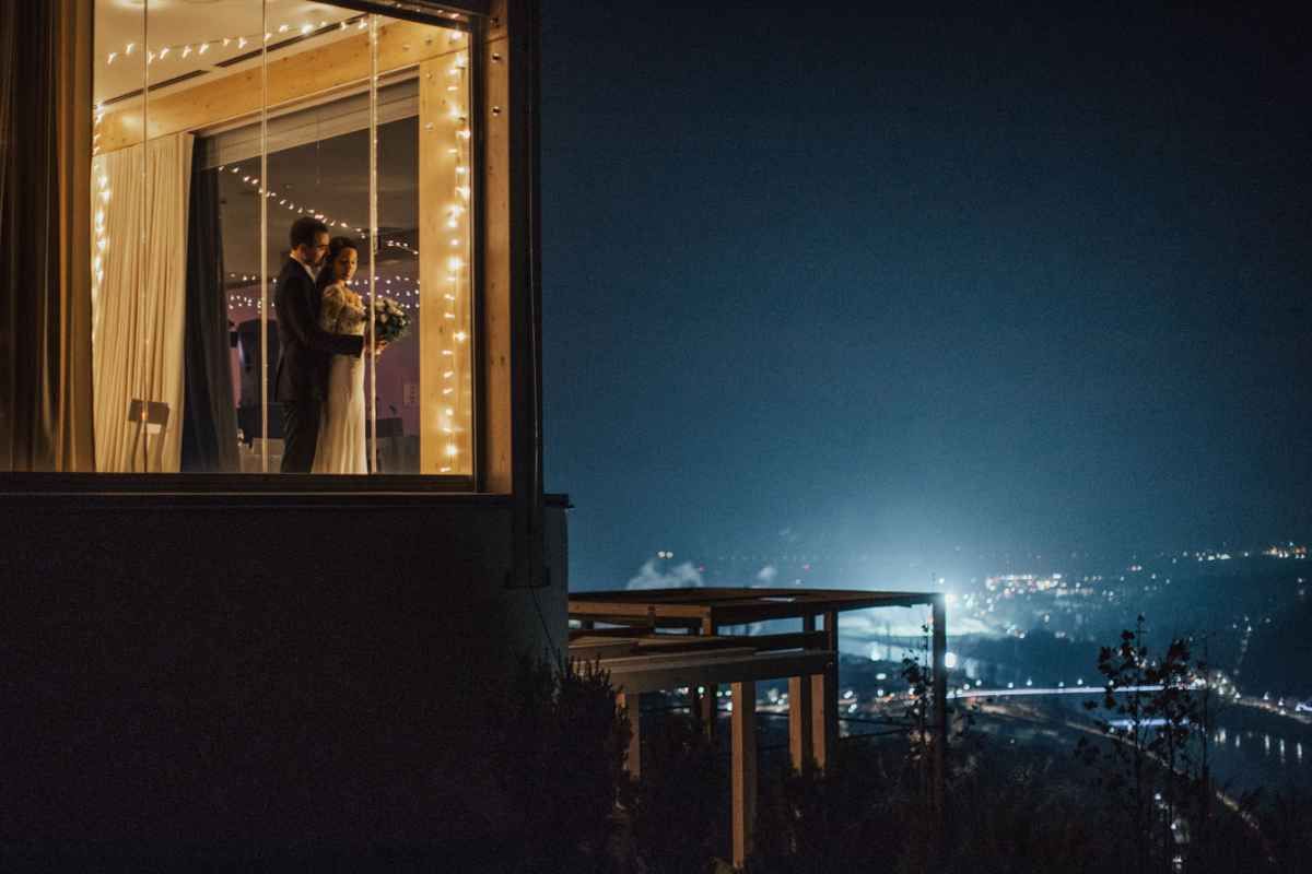 Couple embracing in a warmly lit window at night with city lights glowing in the background.