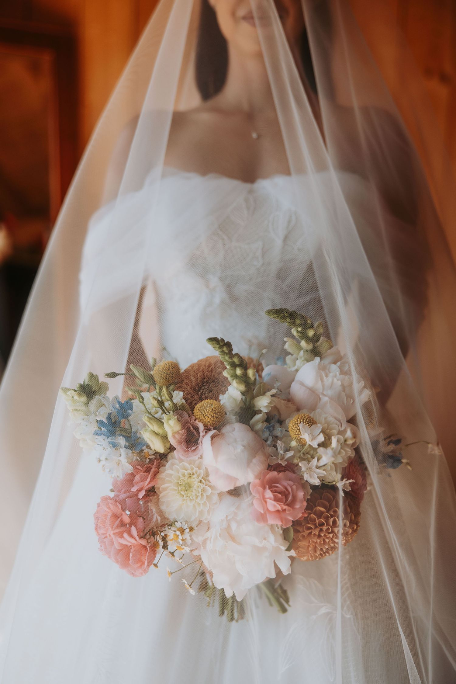 Bride in white lace gown and veil holding a pastel wedding bouquet with pink roses, white peonies, and dried flowers.