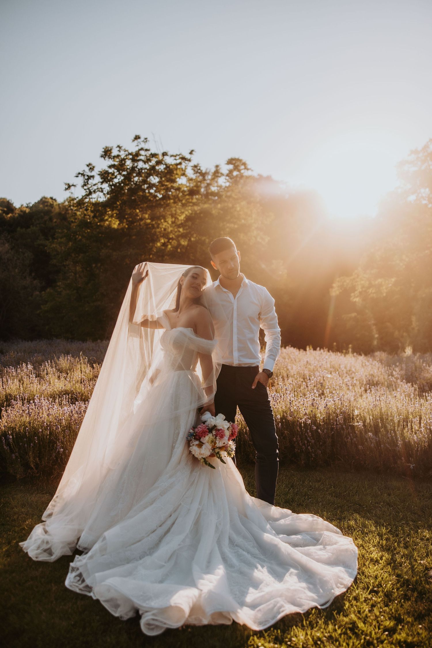 Bride in a long veil and groom stand together in a sunlit lavender field at golden hour.