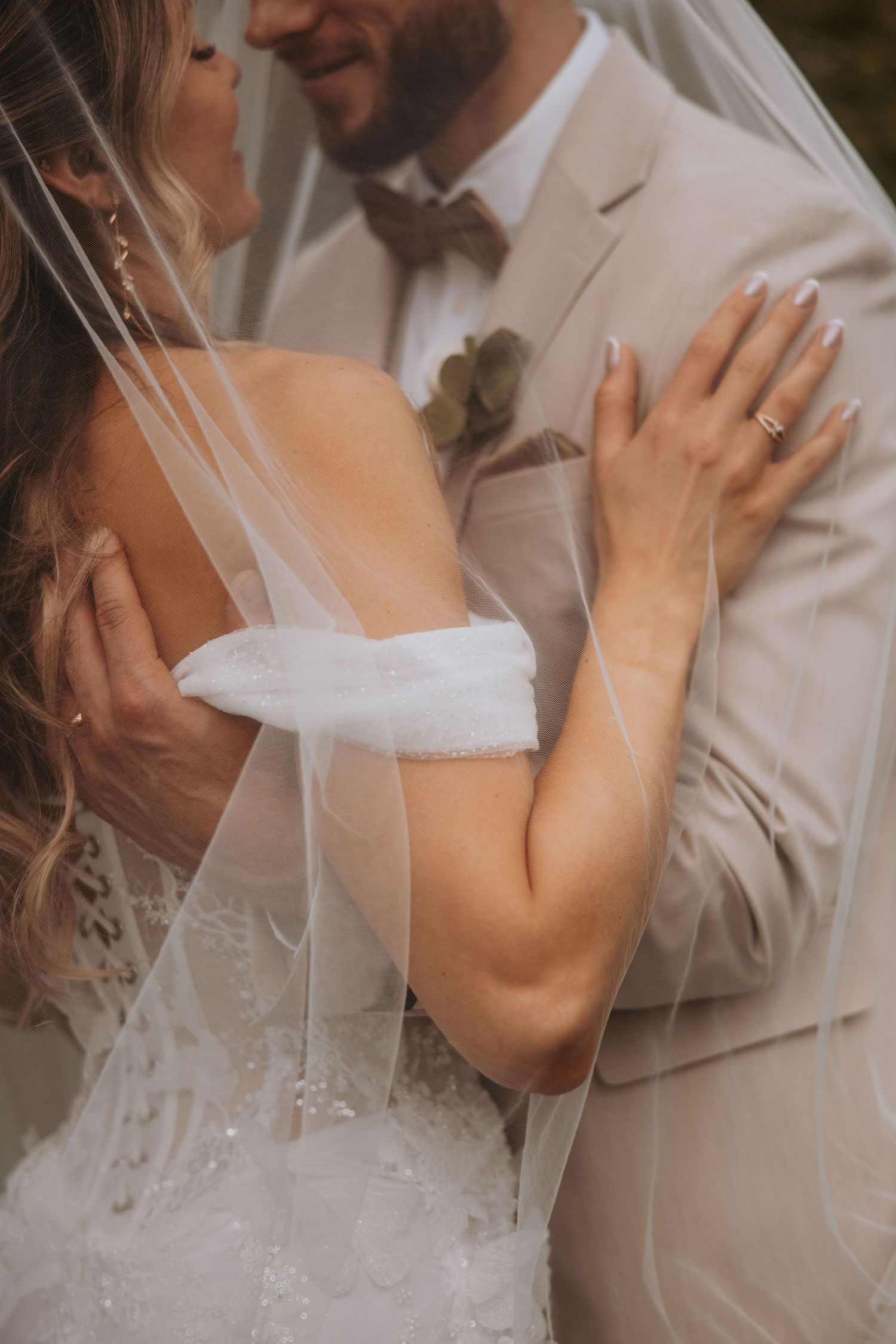 Close-up of bride and groom embracing under veil, bride wearing lace off-shoulder gown with ring.
