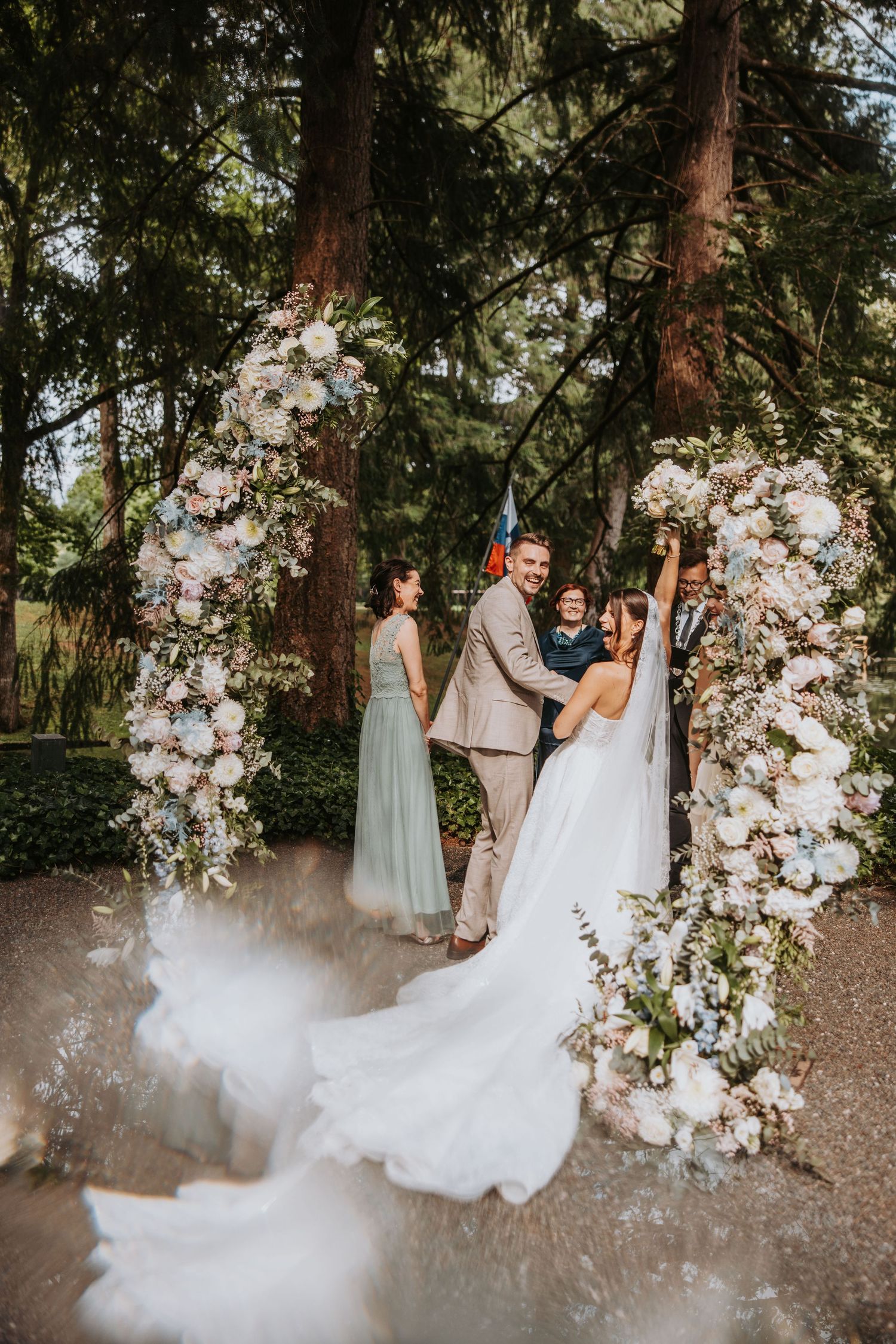 Bride and groom exchange vows under a floral arch surrounded by tall trees, with a bridesmaid and officiant nearby.