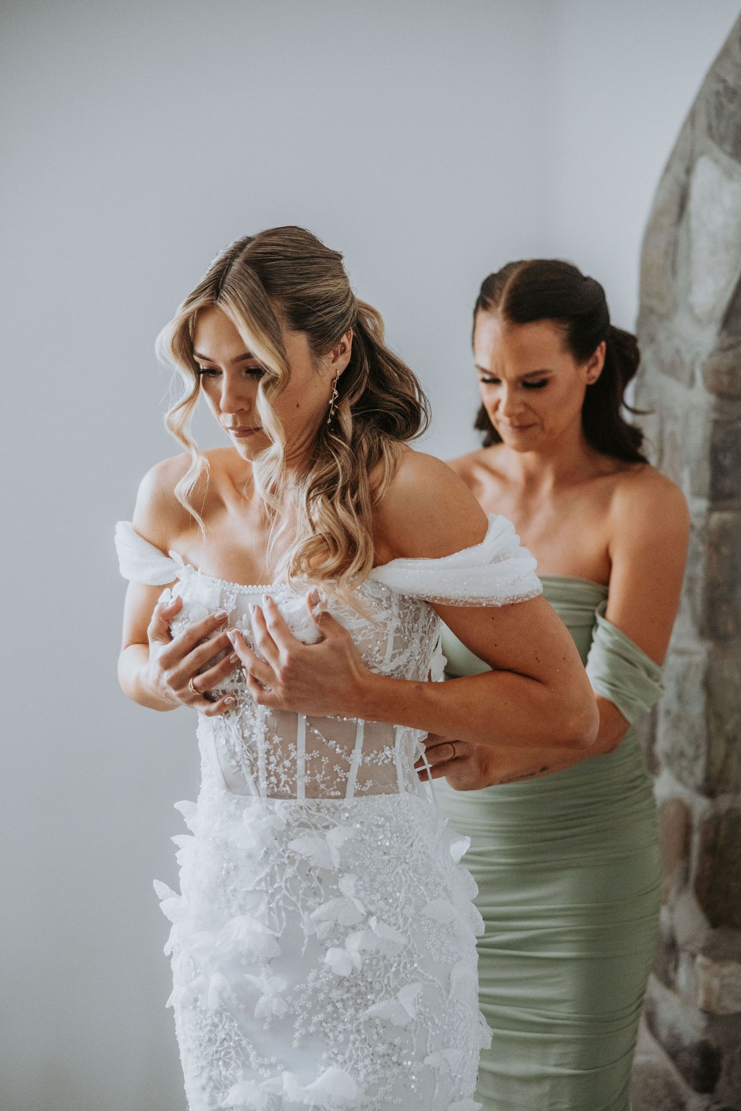Bridesmaid in sage green dress helps bride into off-shoulder lace wedding gown indoors.