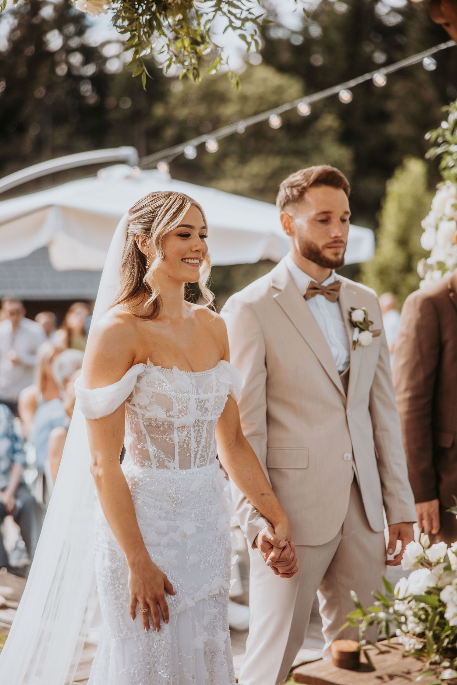 Smiling bride in off-shoulder lace gown walks hand in hand with groom in beige suit at outdoor ceremony.