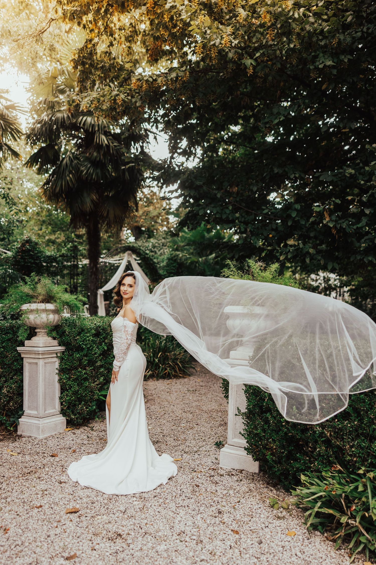 Bride in white gown with flowing veil caught by wind in lush garden setting.