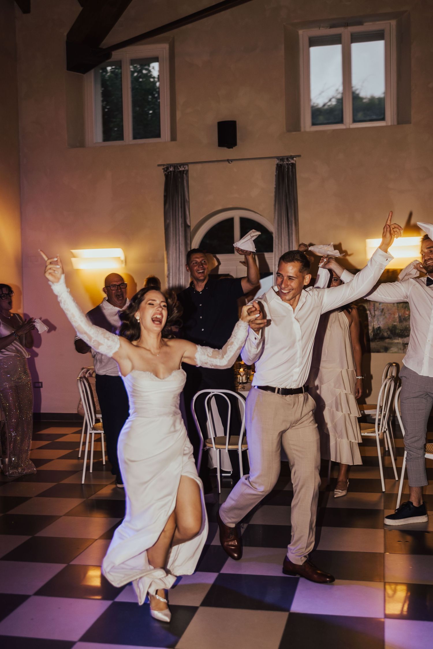 Joyful bride and groom celebrate their first dance entrance with raised arms in a warmly lit wedding reception hall.