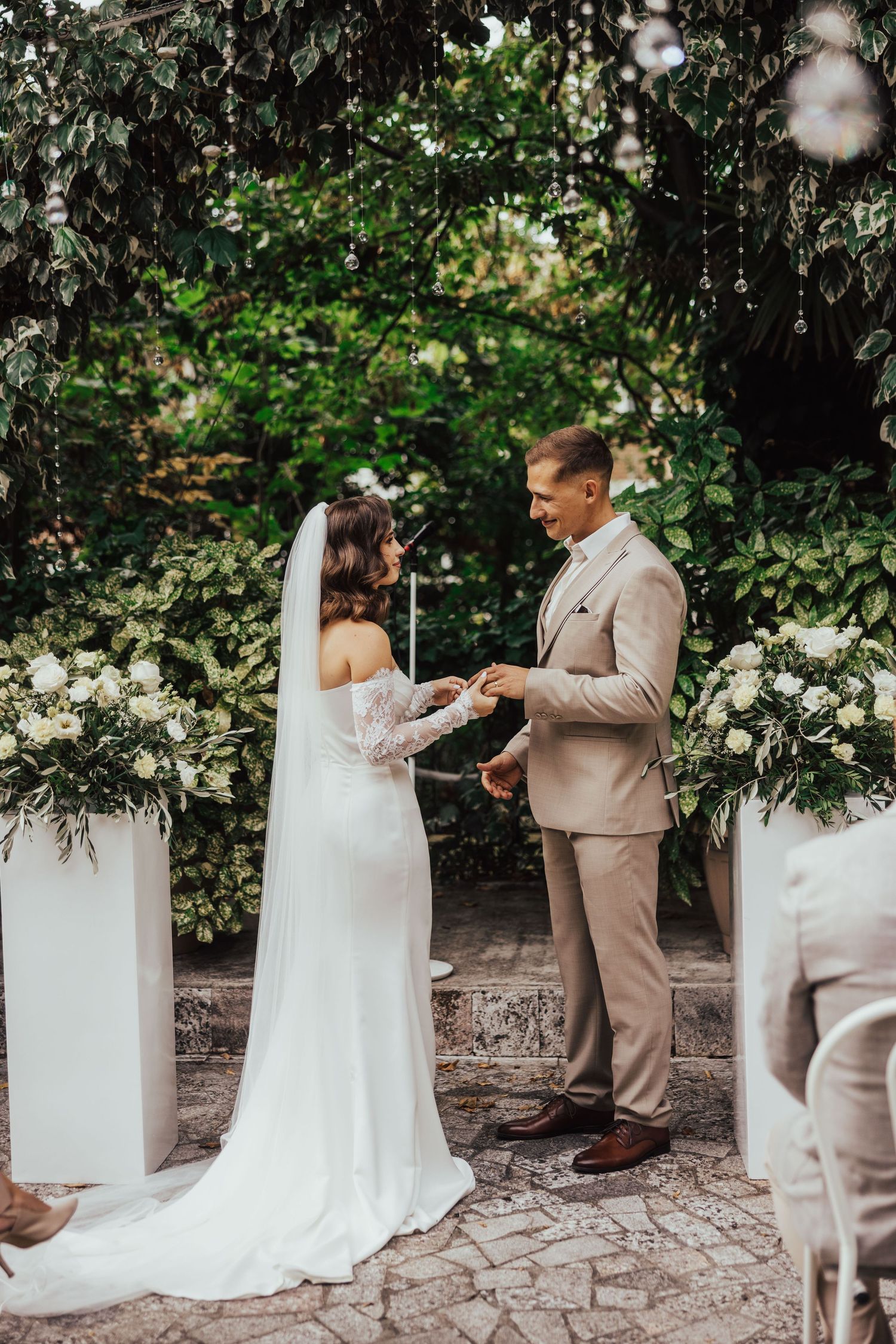 Groom places wedding ring on bride's finger at outdoor garden ceremony with white floral arrangements.