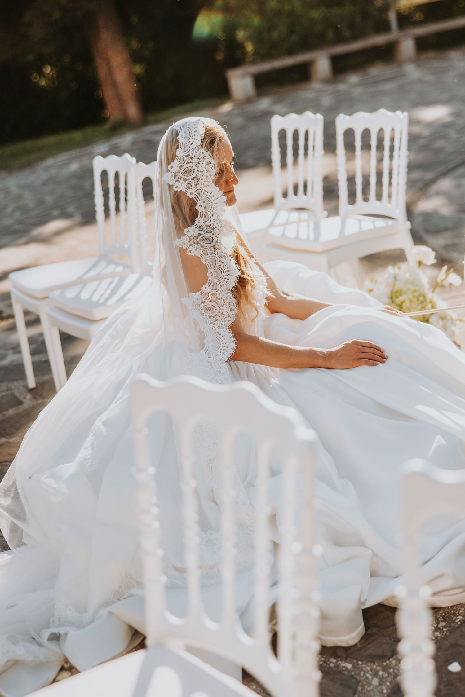 Bride in lace-trimmed veil and ball gown seated among white chairs at outdoor wedding venue.