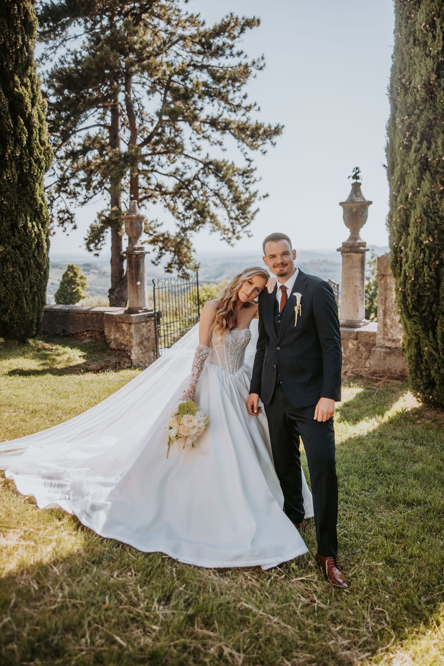 Bride and groom posing in sunlit Tuscan garden with cypress trees and stone gates.