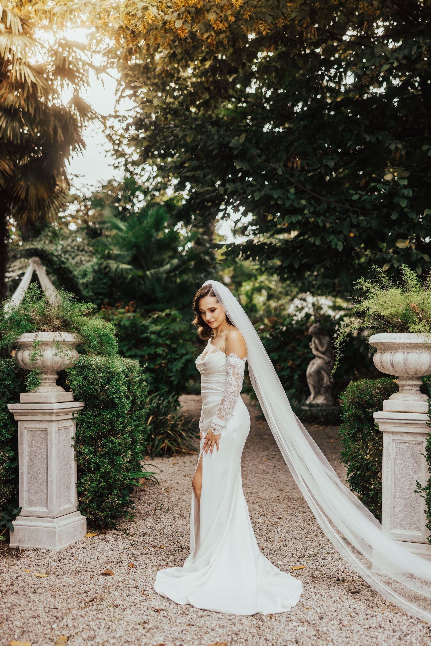 Elegant bride in off-shoulder wedding dress with long veil standing on gravel path in golden garden light.