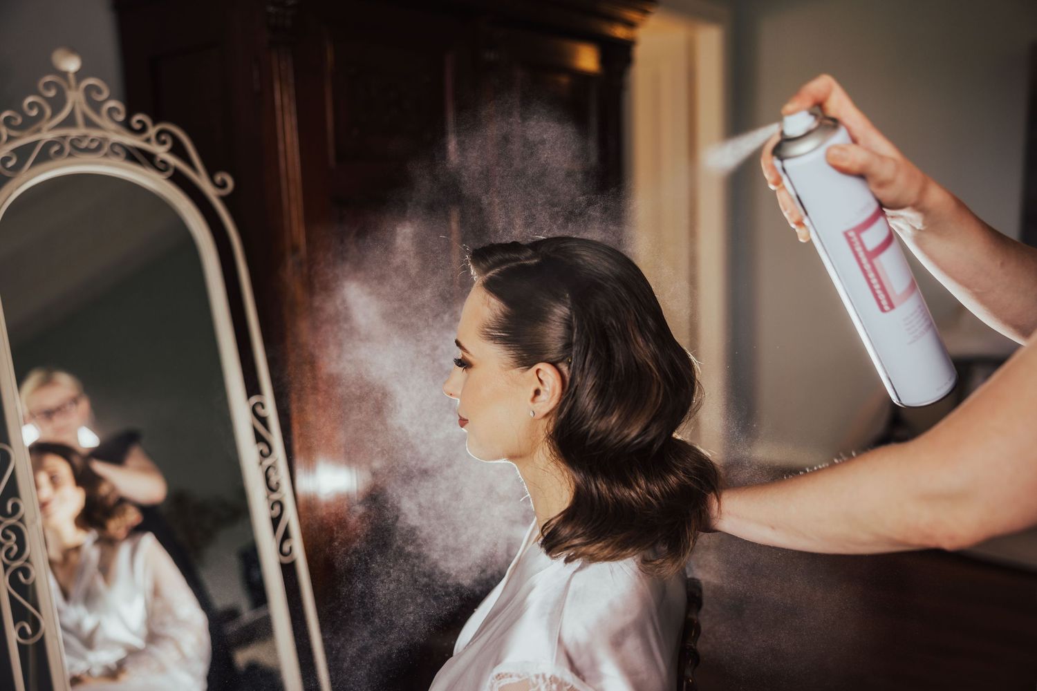 Hairstylist applying hairspray to a brunette woman sitting in front of a mirror during bridal hair preparation.