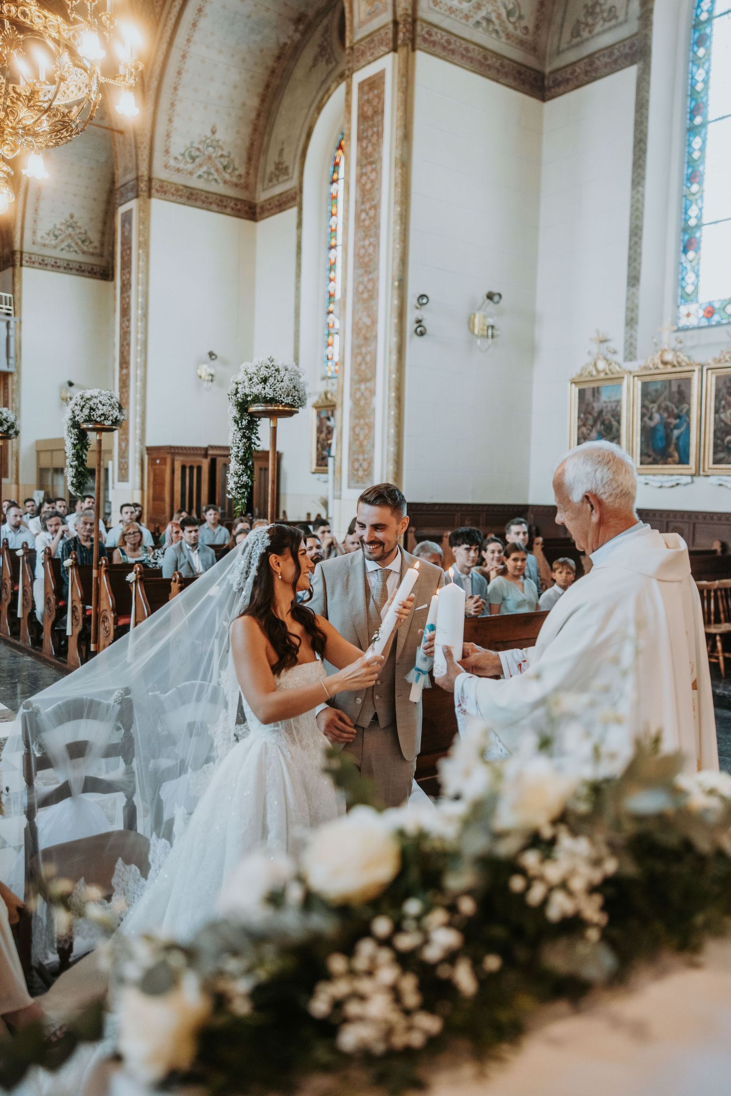 Bride and groom exchange vows at ornate church altar with white floral decorations and guests.