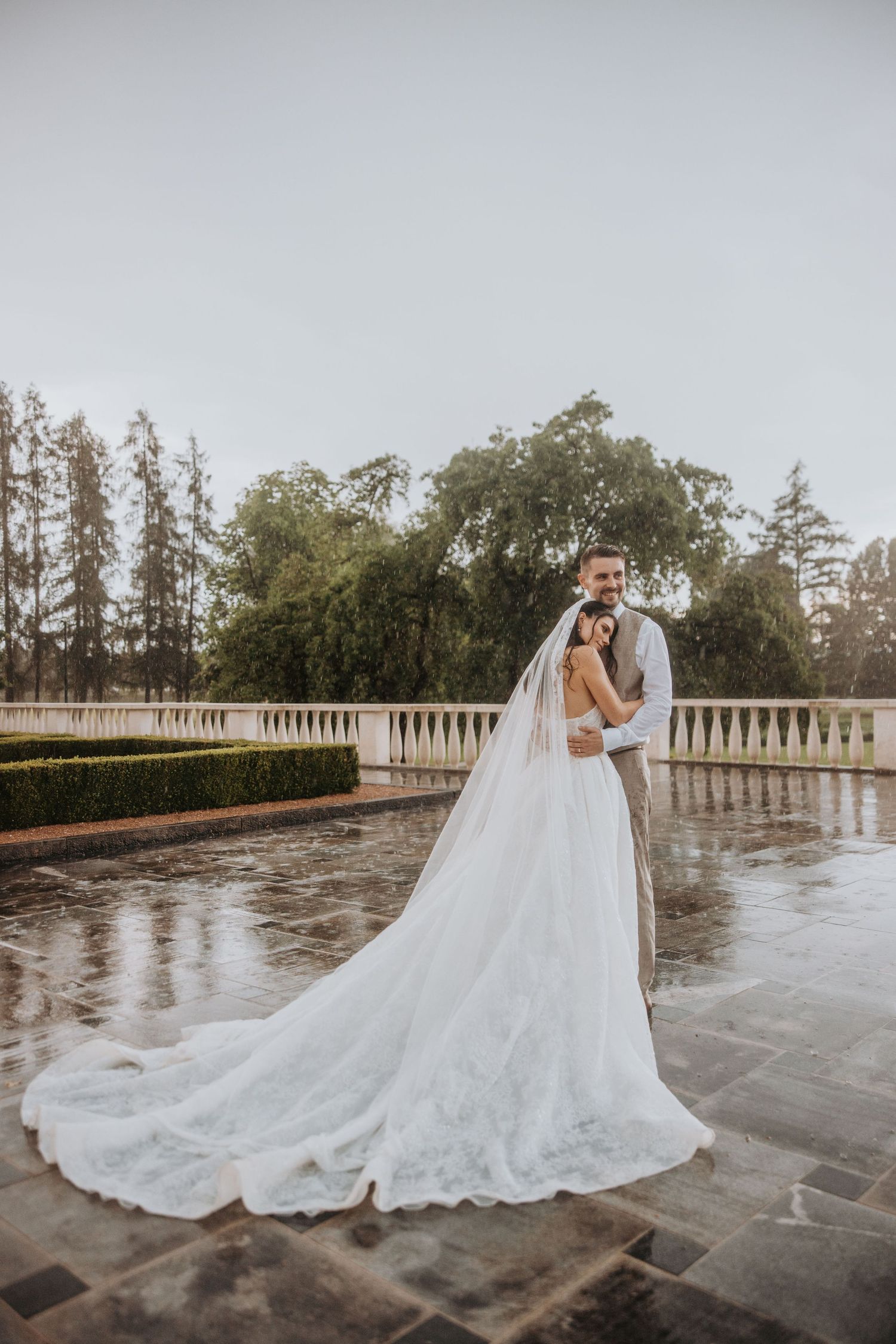 Bride and groom embrace on a rain-soaked terrace, her lace cathedral veil trailing elegantly behind her.