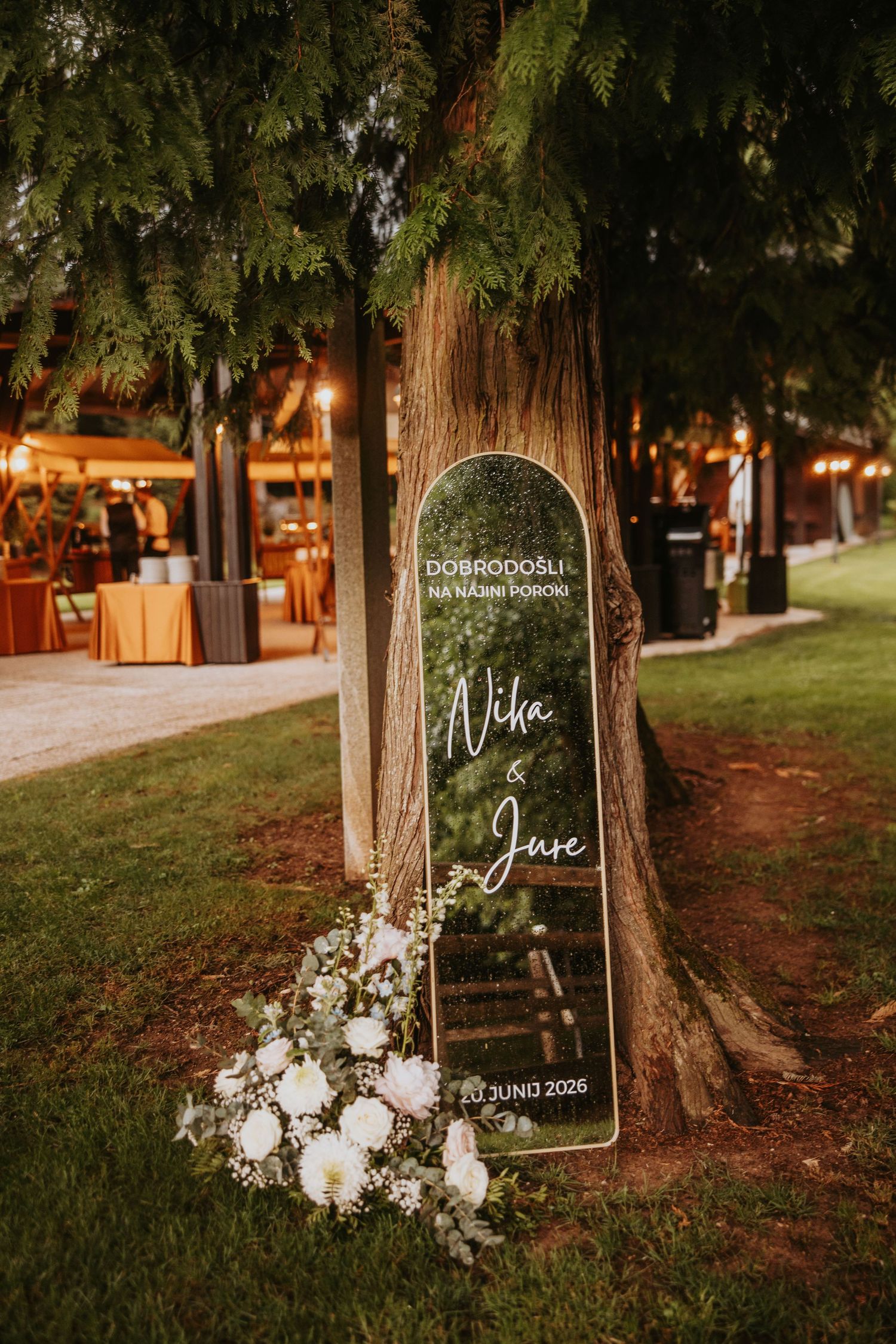 Elegant wedding welcome mirror sign for Nika and Juse leaning against a tree, decorated with white flowers at night.