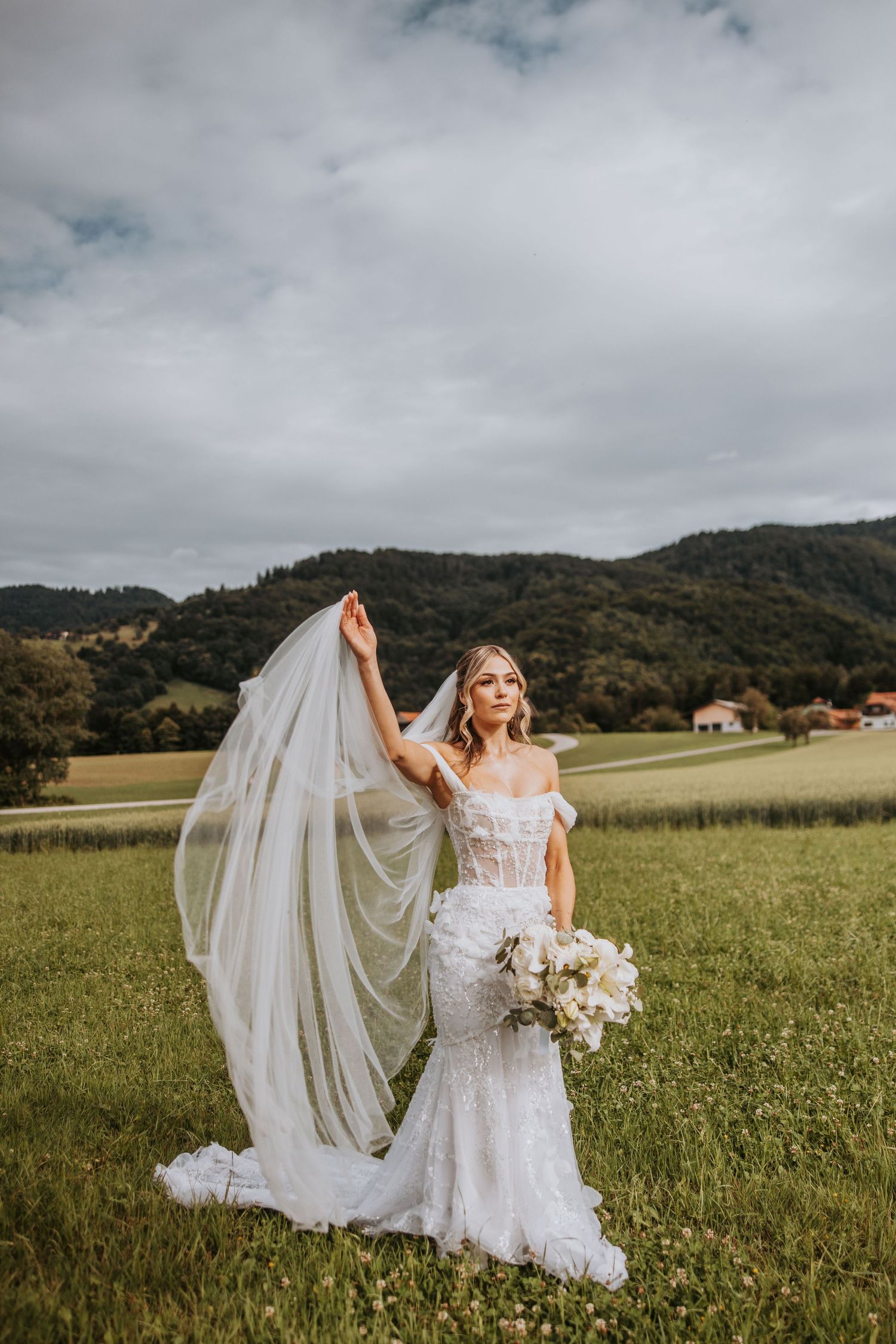Bride in lace wedding dress lifting veil in sunlit field with rolling green hills in background.
