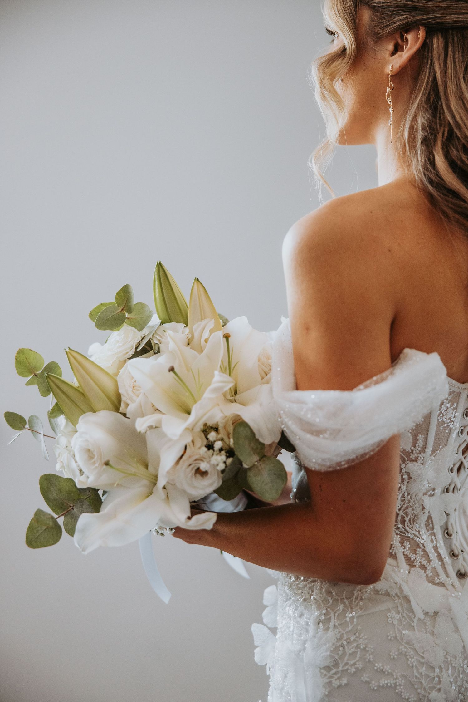 Bride holding elegant white lily and rose bouquet with eucalyptus in off-shoulder lace wedding gown.