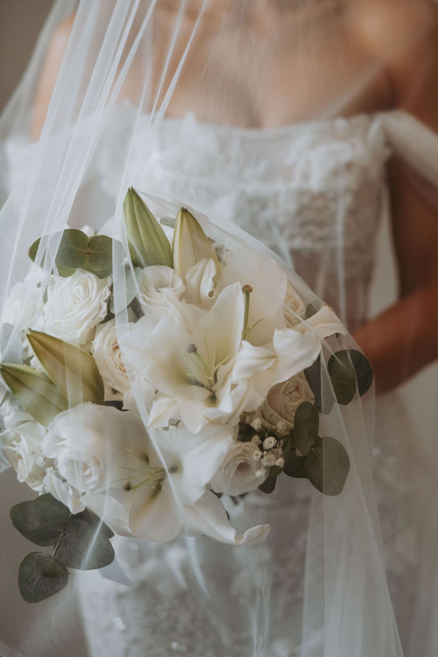 Bride holding elegant white bouquet of lilies and roses draped with flowing veil.