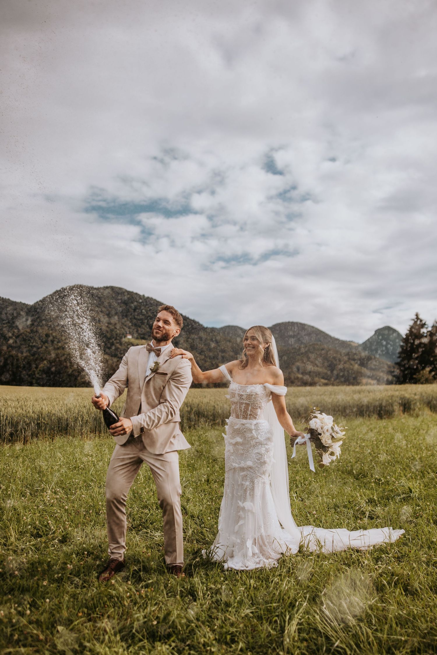 Groom pops champagne bottle in green meadow as laughing bride holds bouquet with mountain views behind.