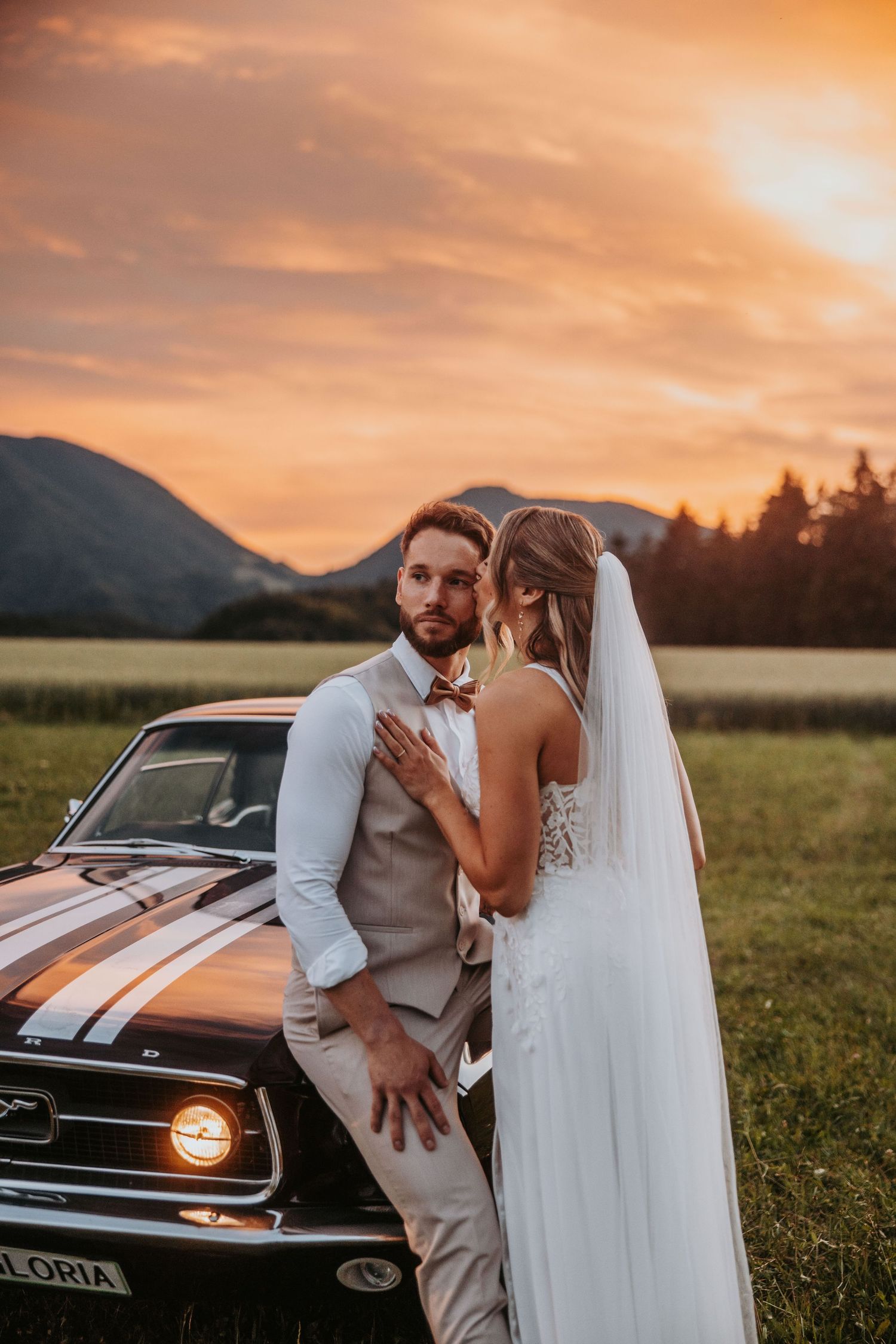 Groom in beige suit embraces bride in lace gown beside classic Mustang at vibrant sunset.