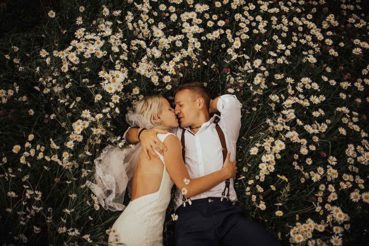 Romantic bride and groom lying together in field of white daisies during wedding photo session.