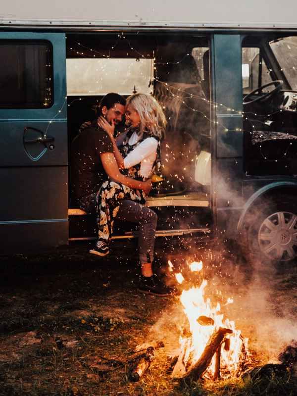Couple embracing romantically in open van doors with a glowing campfire in the foreground at dusk.