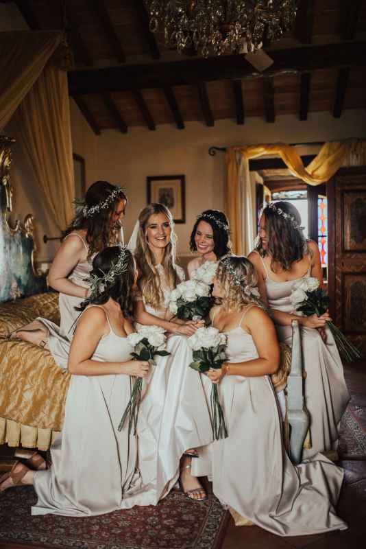 Bridesmaids in white satin gowns with floral crowns gather around bride holding white roses in elegant rustic villa.