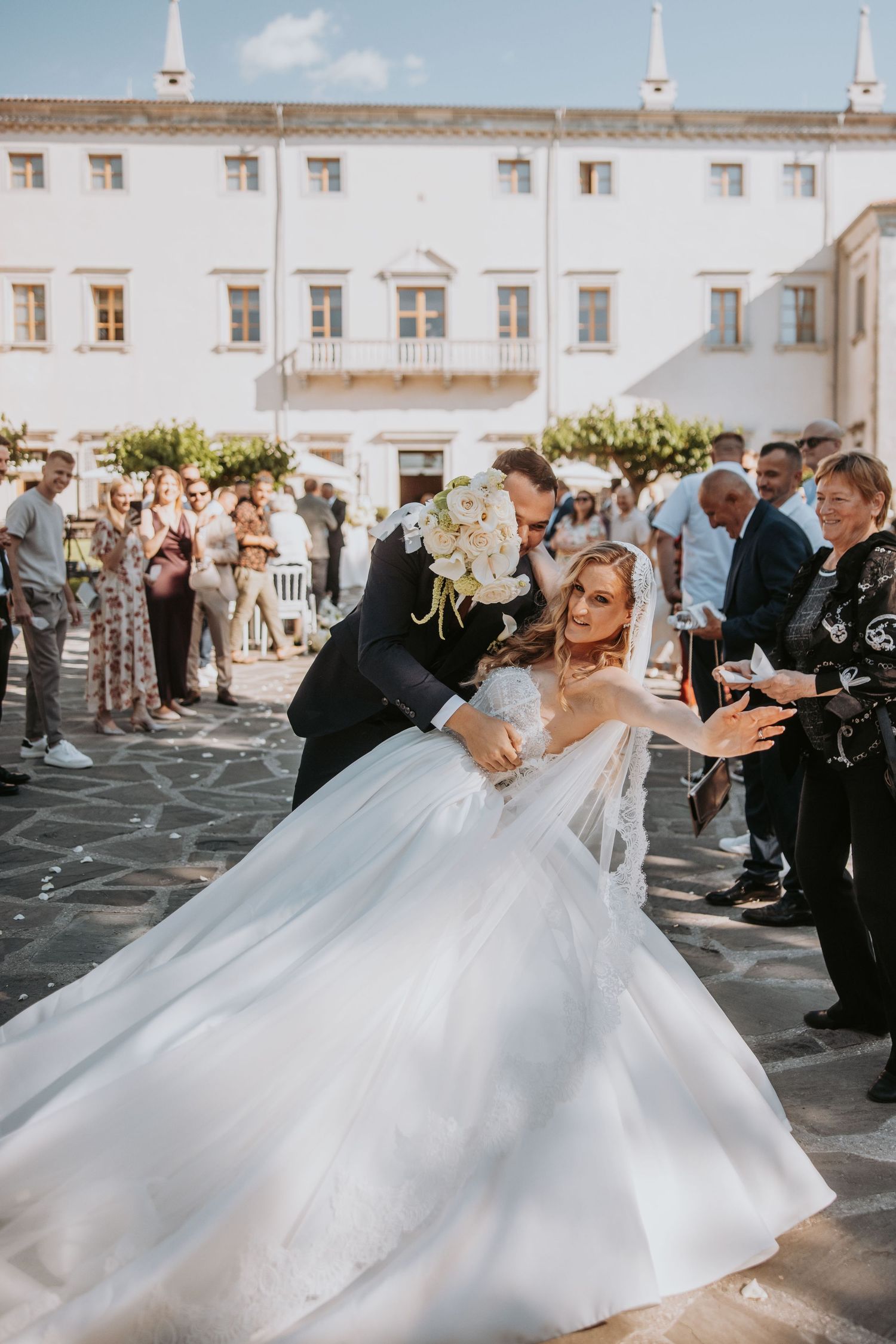 Bride and groom share a romantic dip kiss outside a white venue while wedding guests celebrate around them.