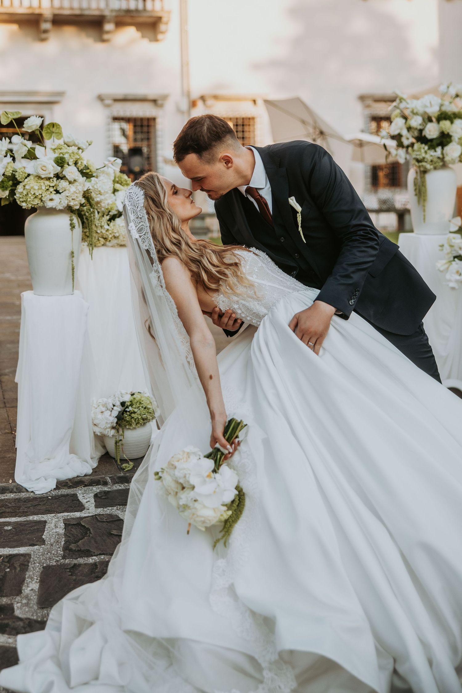 Groom dips bride for romantic kiss outdoors surrounded by elegant white floral arrangements at wedding.