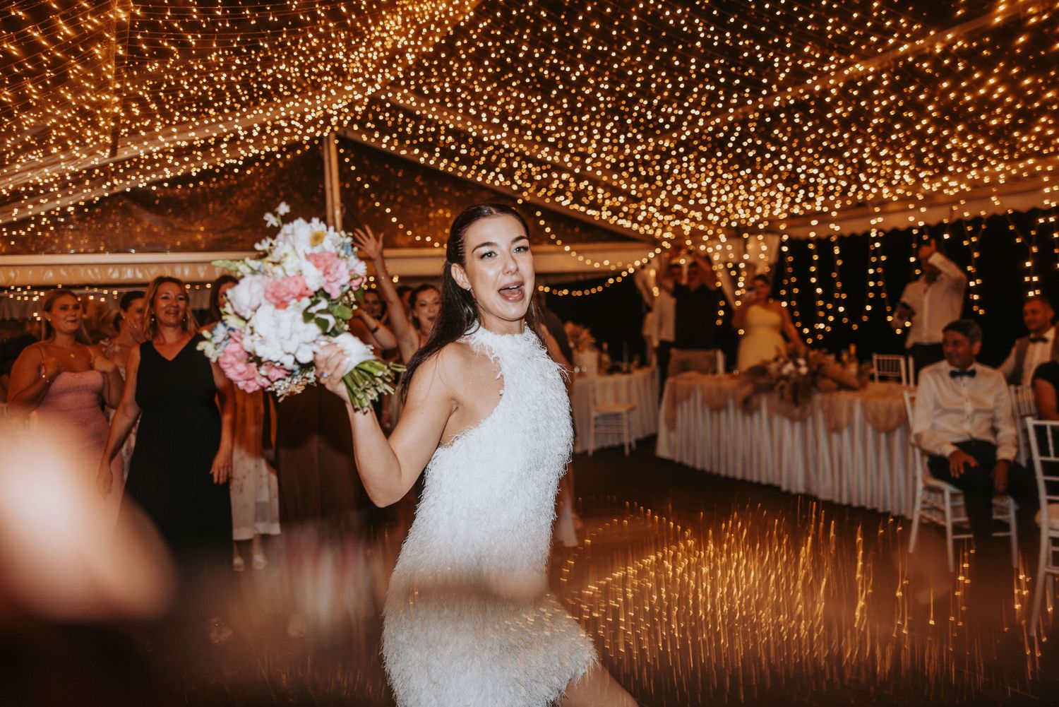 Bride in white dress joyfully tossing bouquet on glowing dance floor under twinkling fairy lights at wedding reception.