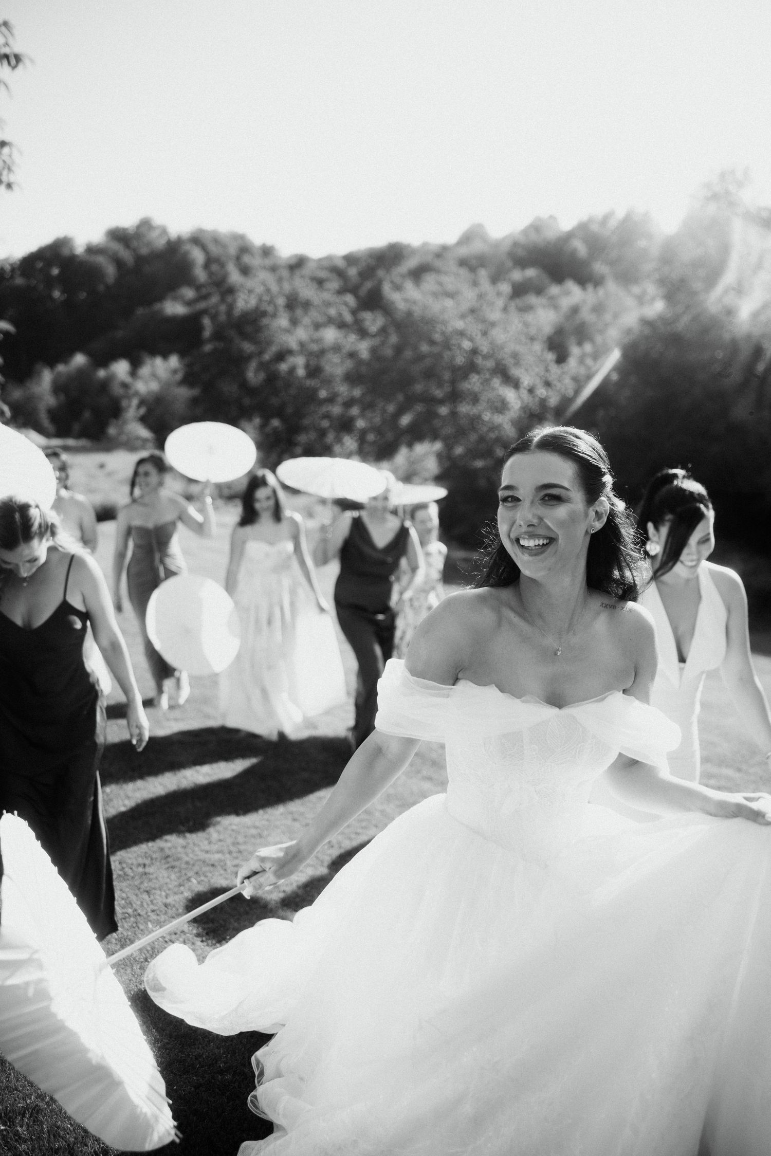 Black and white photo of a smiling bride in an off-shoulder gown walking with bridesmaids holding parasols.