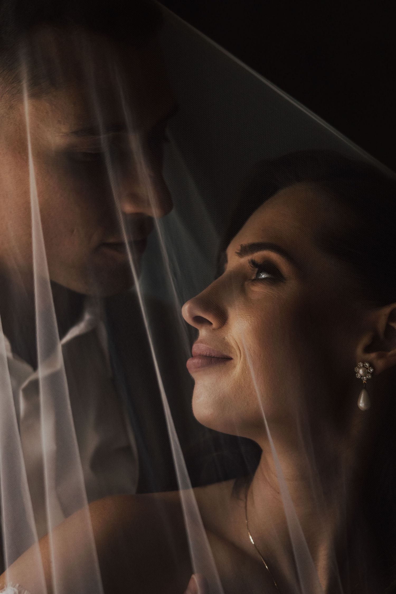 Intimate close-up portrait of bride and groom under sheer wedding veil in dramatic lighting.