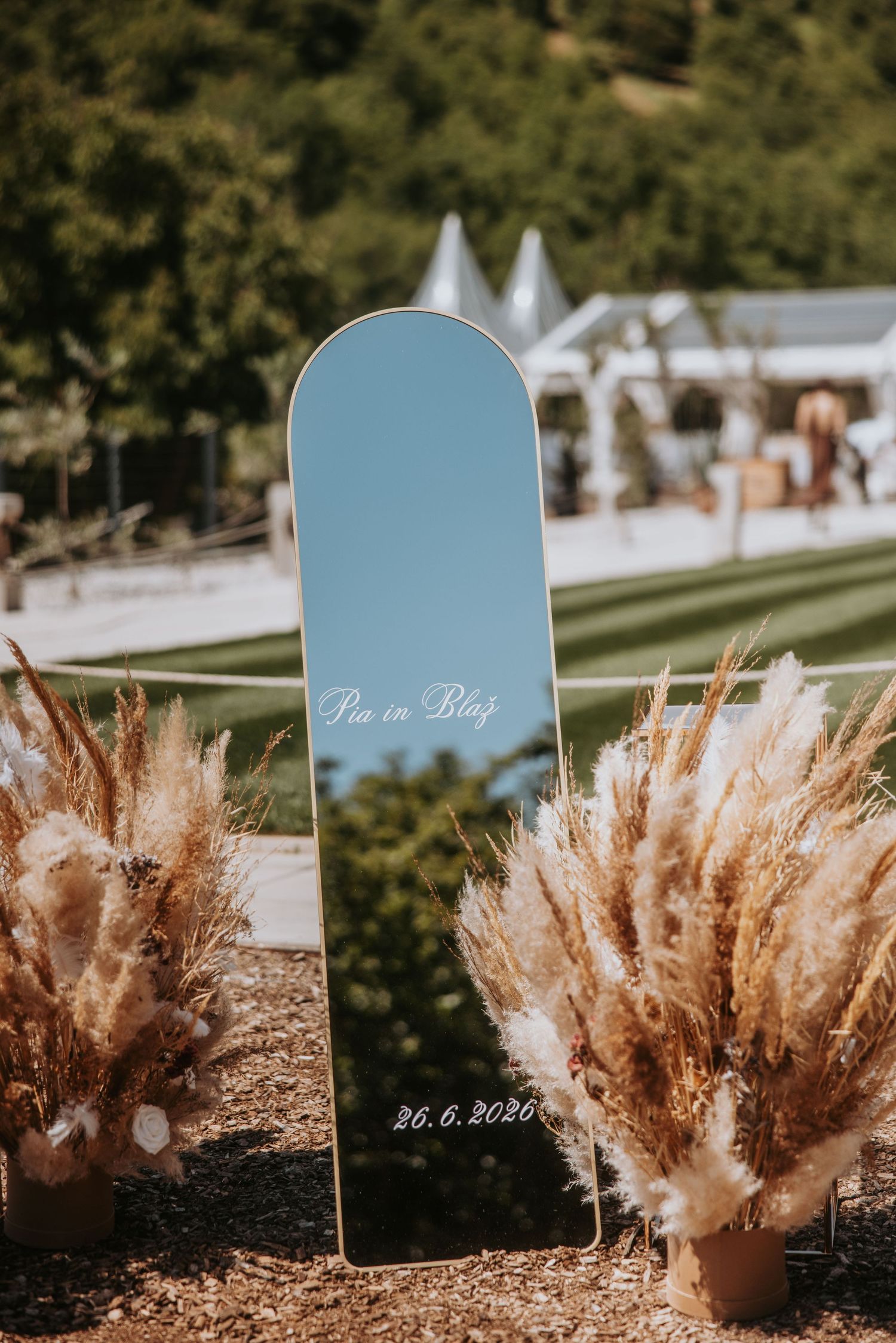 Elegant wedding welcome mirror sign with pampas grass decor reading Pia and Blag dated 26.6.2026 outdoors.