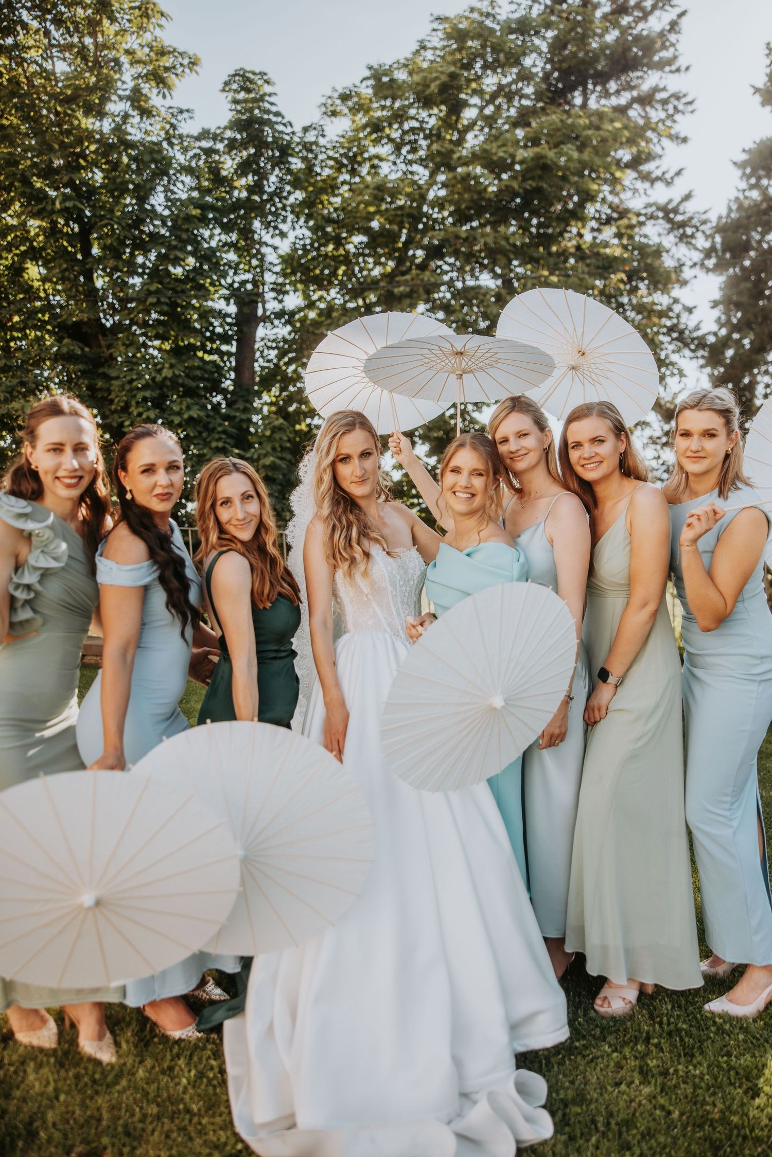 Bride poses with bridesmaids in mint dresses holding white parasols outdoors on a sunny wedding day.
