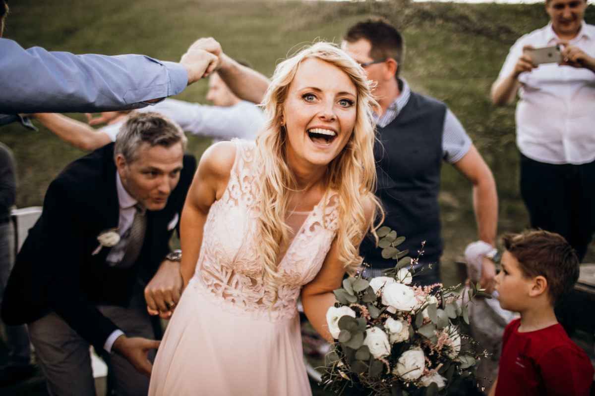 Joyful bride in blush lace gown laughing and holding bouquet surrounded by wedding guests outdoors.
