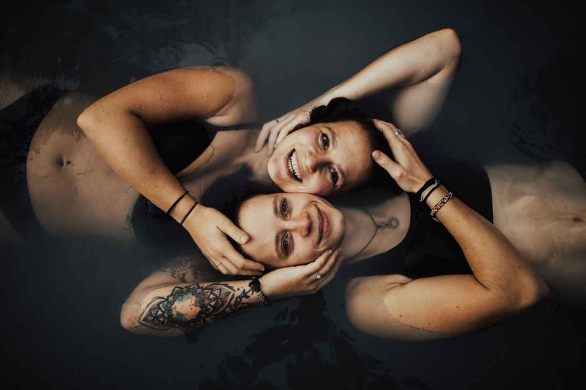 Two people with tattoos lying back-to-back in dark water, smiling in an artistic couples portrait photo.
