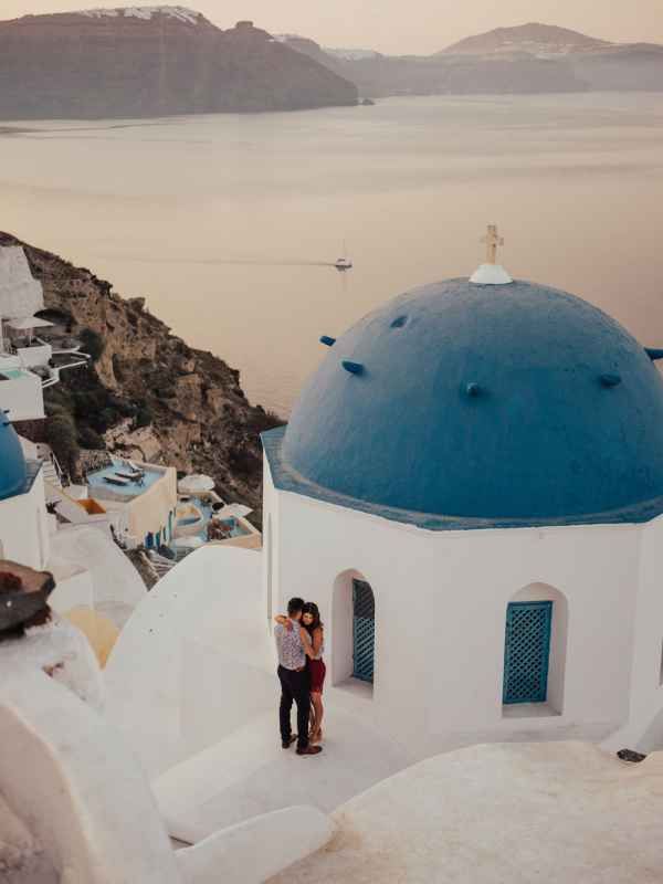 Couple embracing by iconic blue-domed church in Santorini, Greece, with stunning caldera views at sunset.