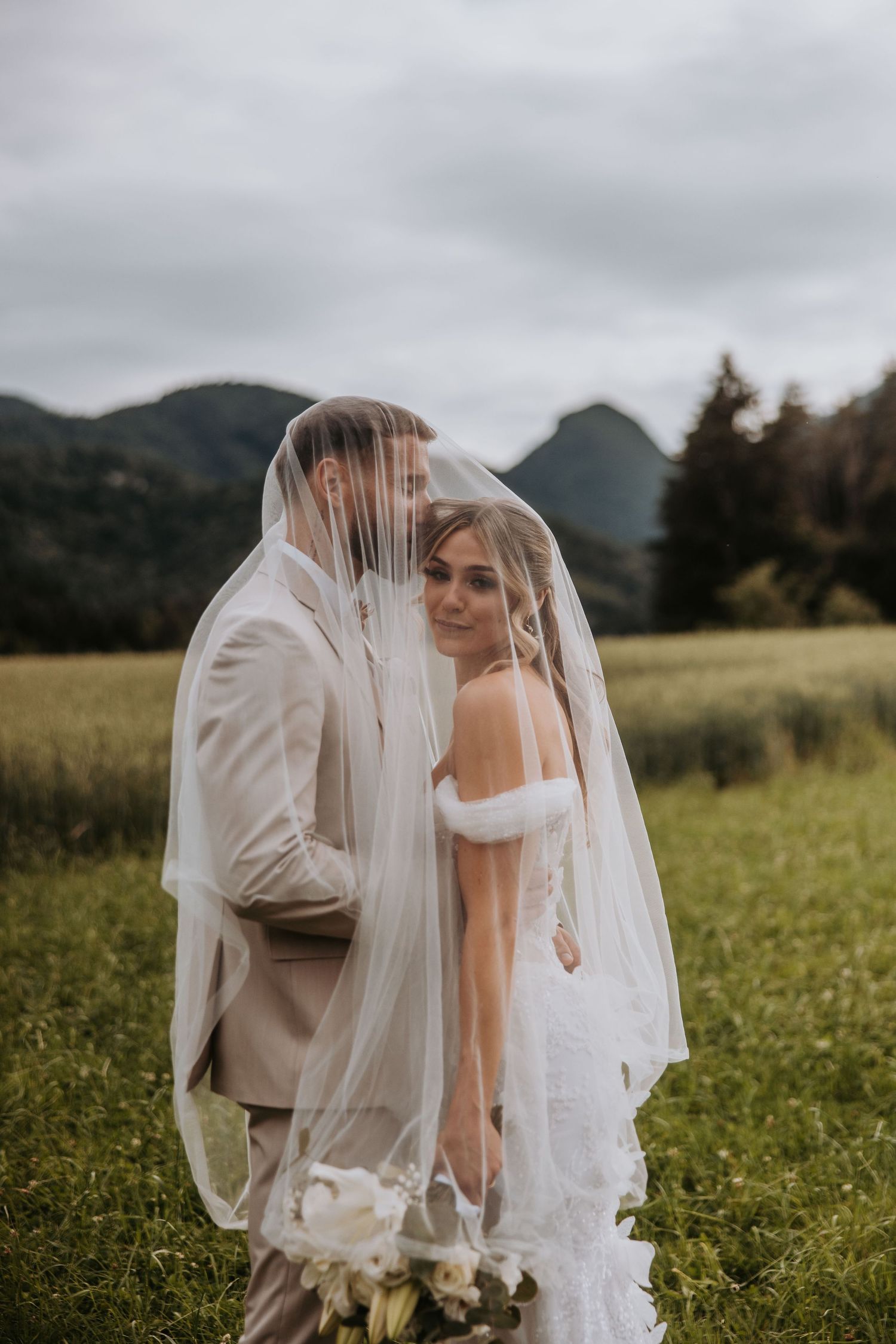 Couple sharing romantic moment under bridal veil in scenic mountain meadow on wedding day.
