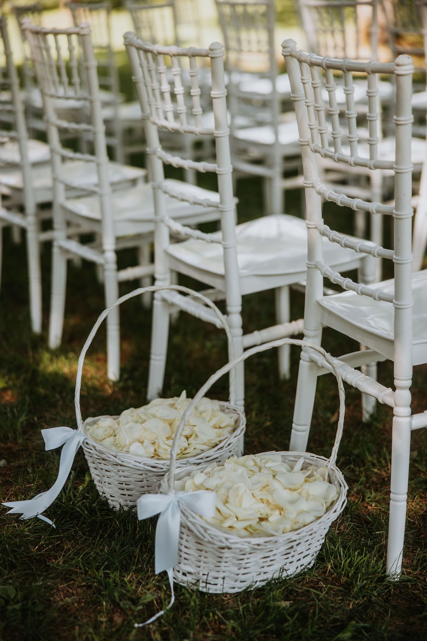 Two white wicker baskets filled with rose petals beside white Chiavari chairs on grass.