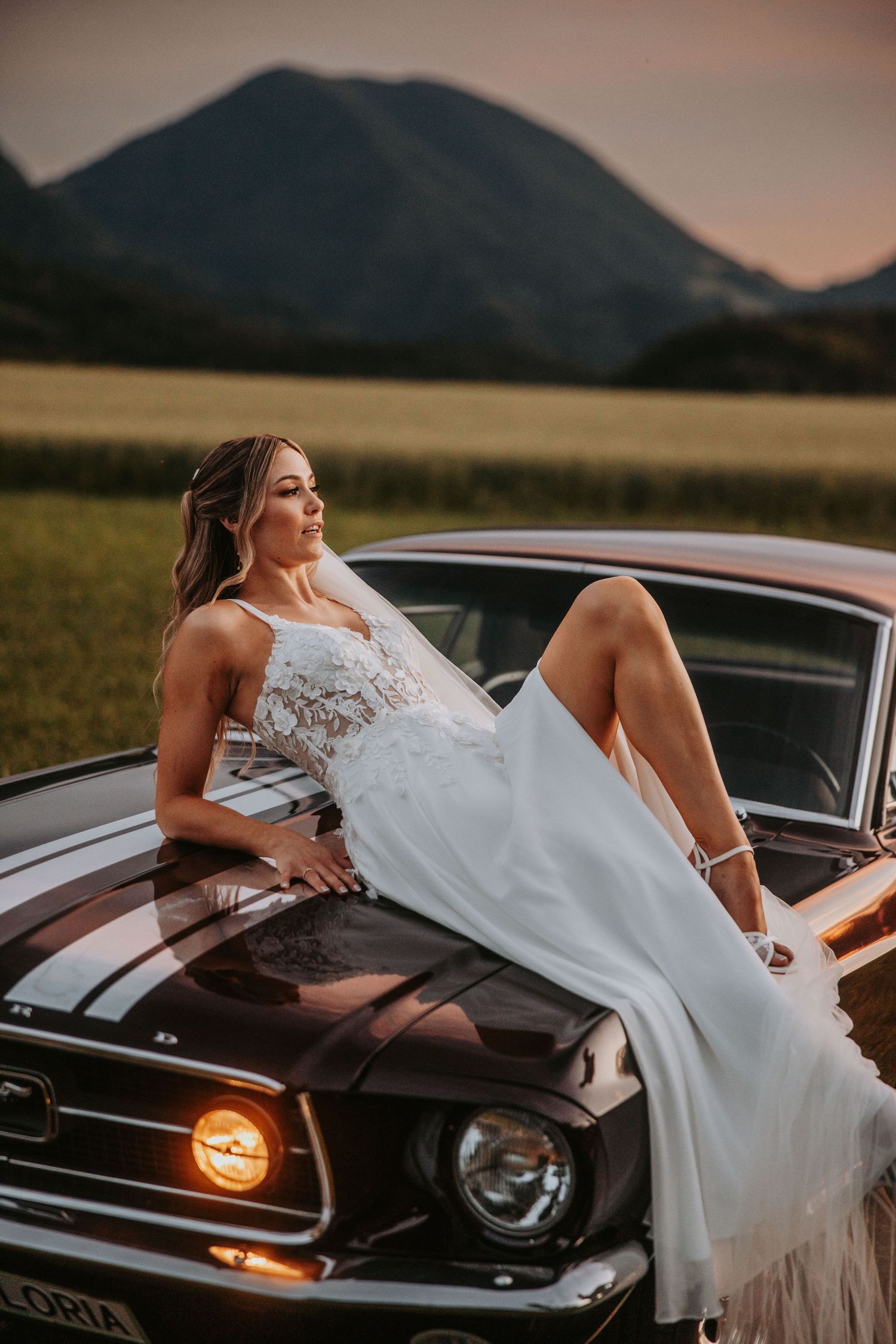 Bride in white lace gown posing on vintage Ford Mustang hood at sunset with mountain views.
