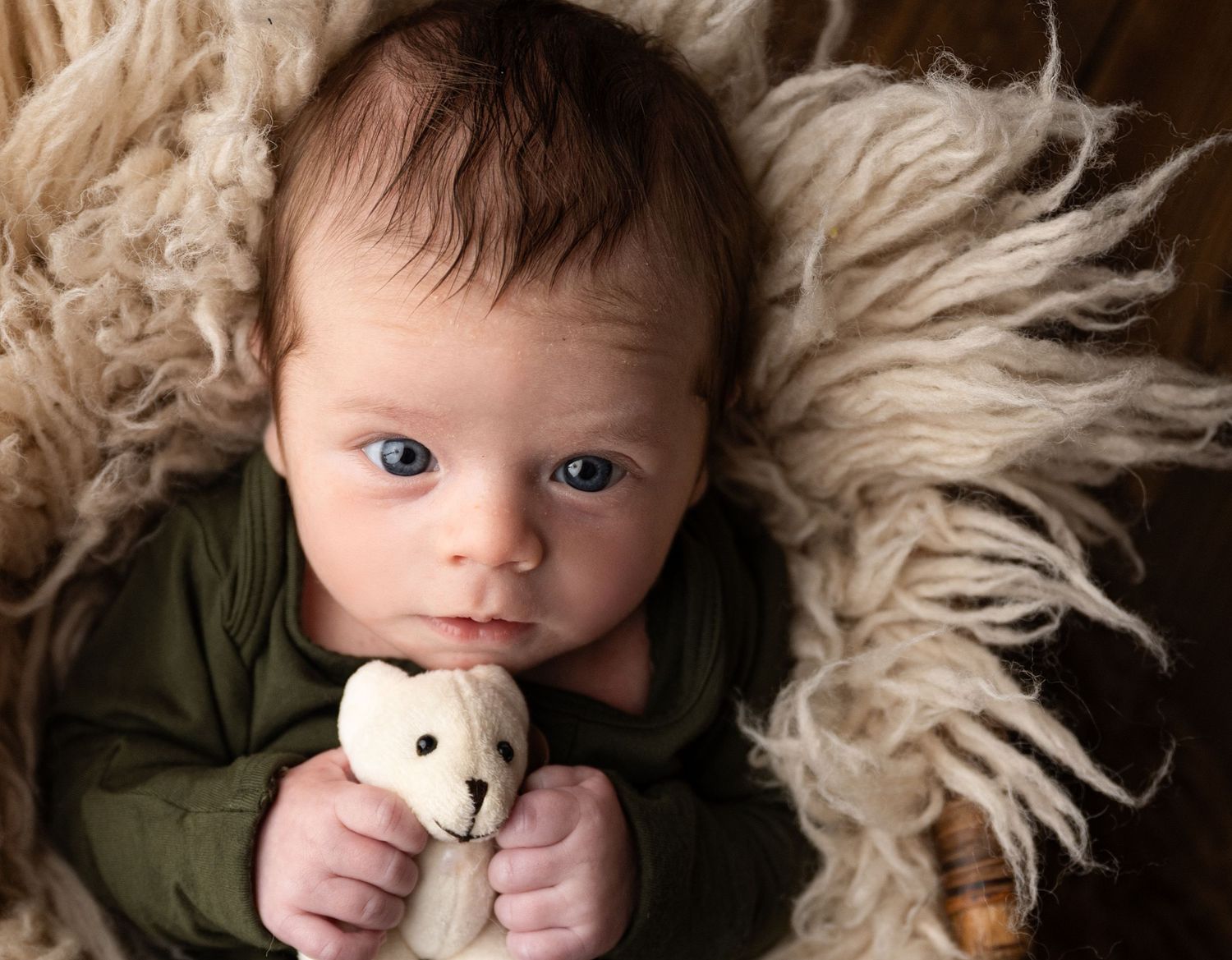 Newborn baby in green onesie holding a tiny white teddy bear, lying on fluffy beige fur with bright blue eyes.
