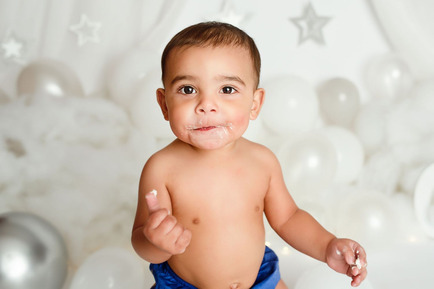 Cute baby boy with cake on face sitting among white balloons at first birthday smash cake photo session.