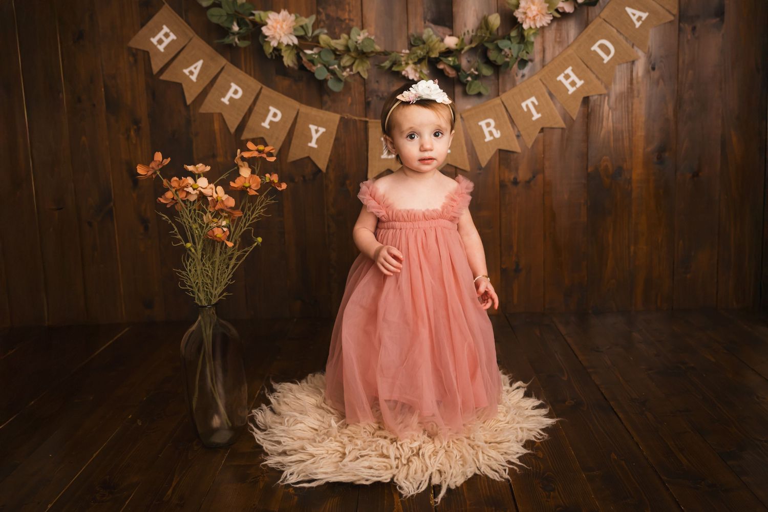 Toddler girl in pink dress stands on fur rug under Happy Birthday banner with floral decor on wood backdrop.