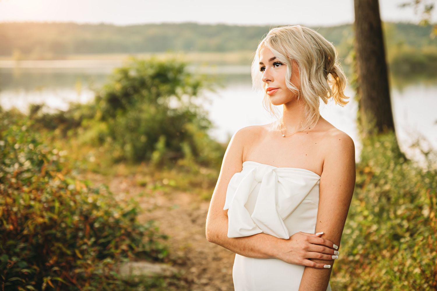 Elegant blonde bride in a strapless white bow gown poses near a serene lakeside at golden hour.