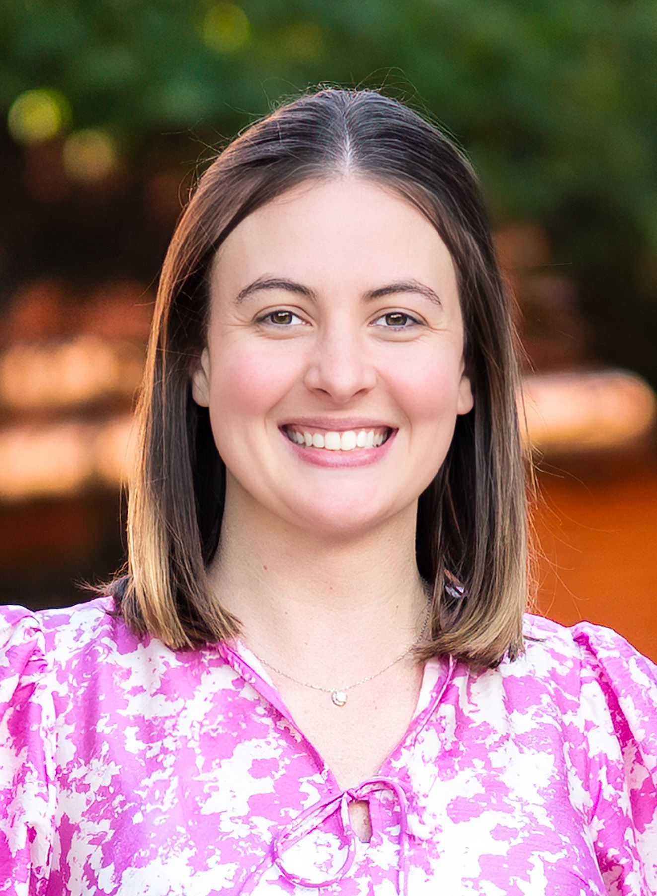 Smiling young woman with brown hair wearing a pink floral blouse, professional headshot with bokeh background.