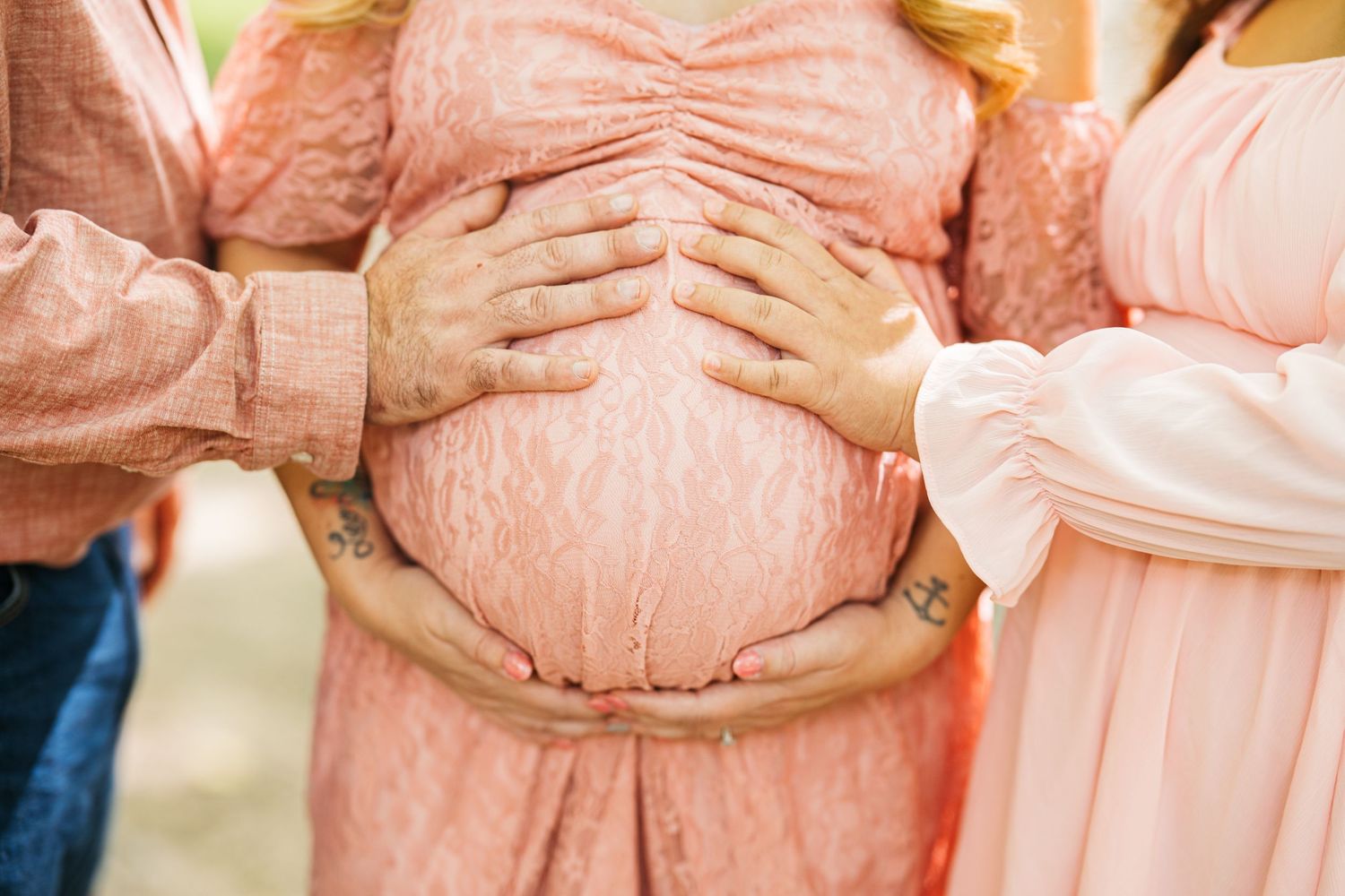 Pregnant woman in pink lace dress with hands of loved ones cradling her baby bump in a maternity photo.
