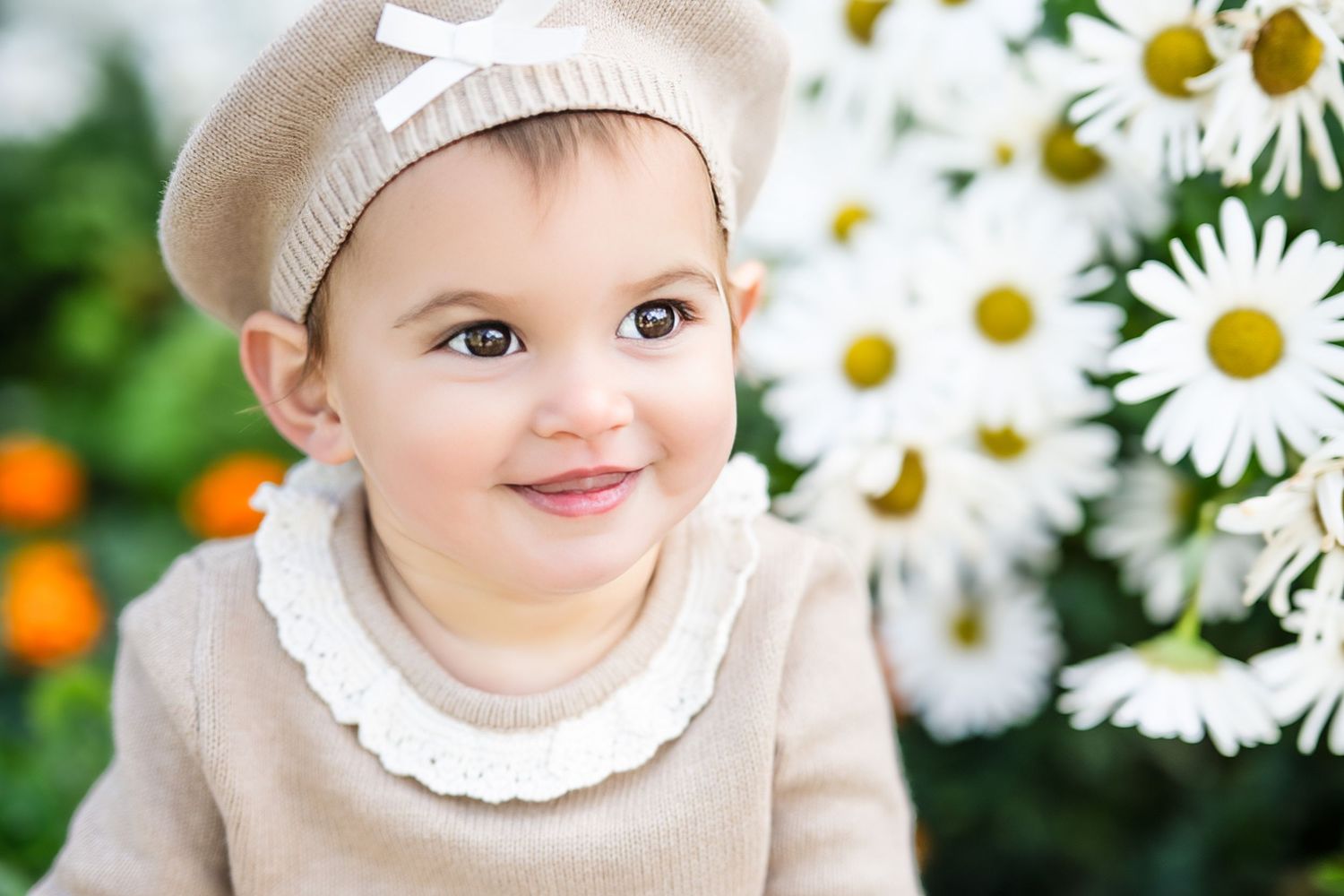 A smiling baby in a beige knit outfit and beret hat poses near white daisy flowers outdoors.